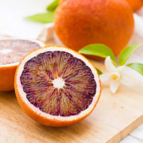 Fresh blood oranges sliced open, showcasing vibrant red pulp, surrounded by green leaves and white flowers.